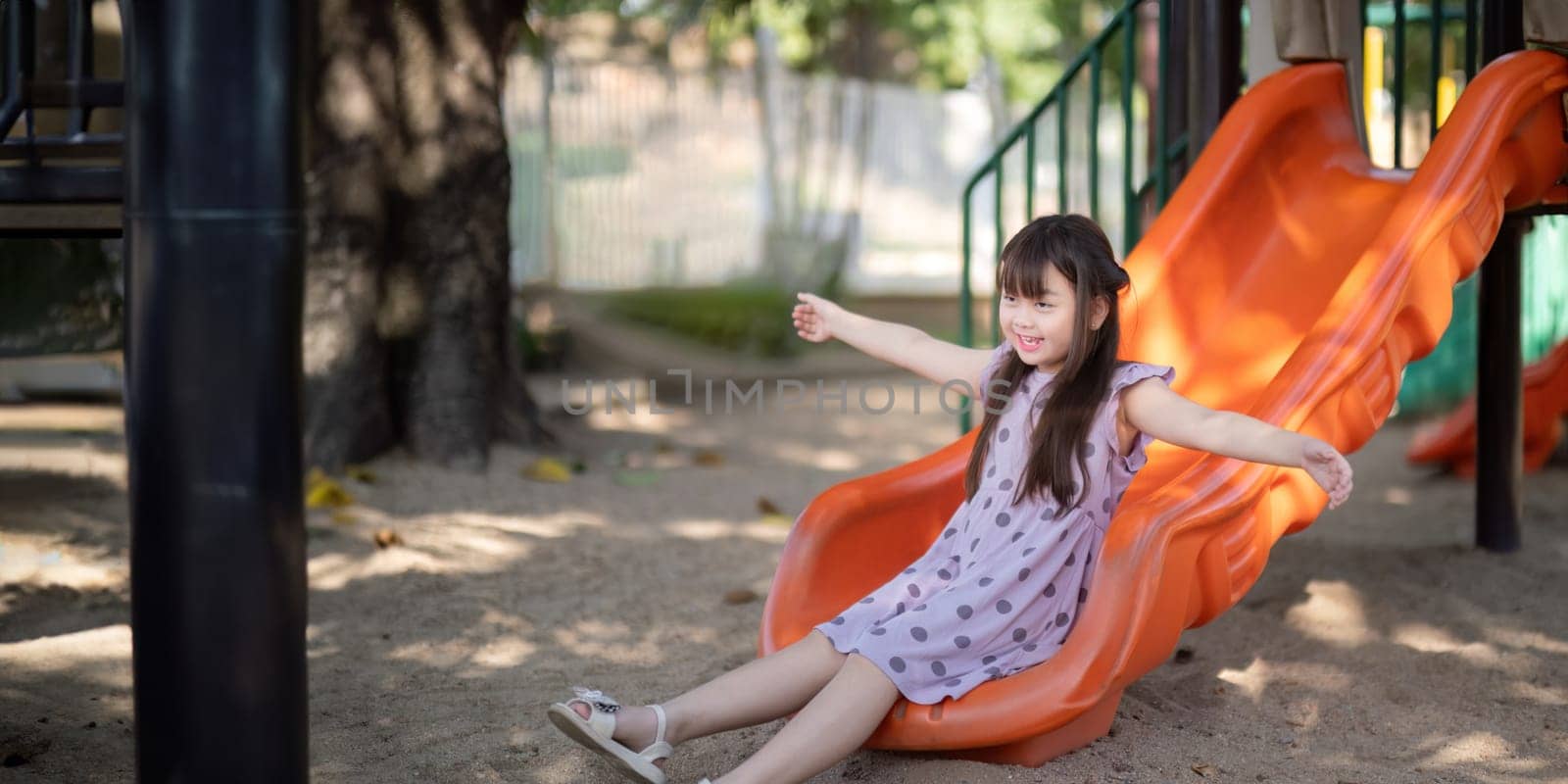 Smiling Asian little girl with fun sitting on slider at playground in the park. Education activity outdoor concept.