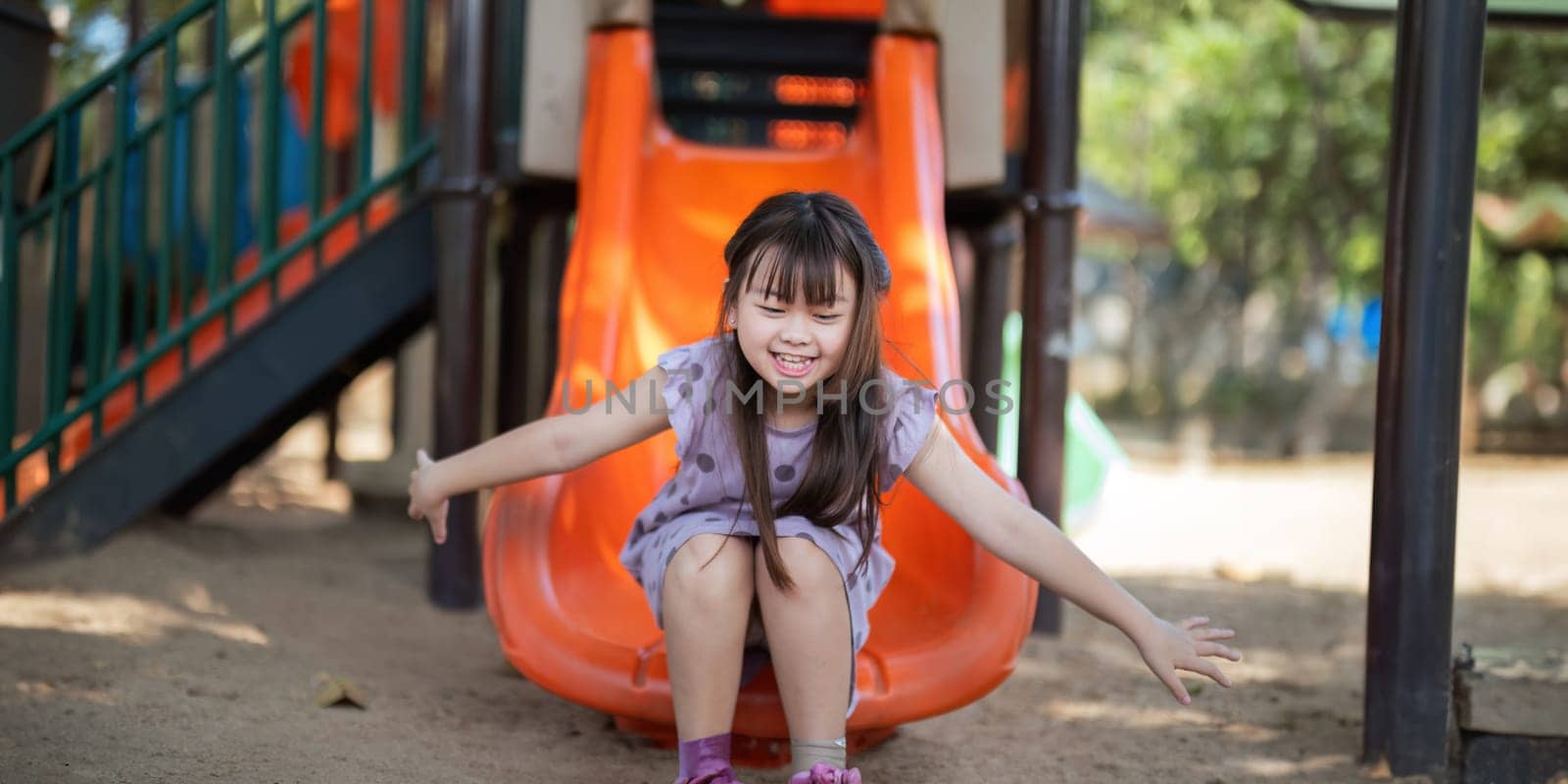 Smiling Asian little girl with fun sitting on slider at playground in the park. Education activity outdoor concept by nateemee