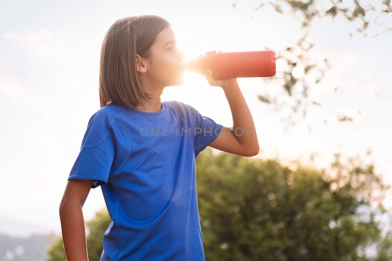 girl drinking water during her training, concept of sport in nature for children and healthy lifestyle, sunlight with flare effect