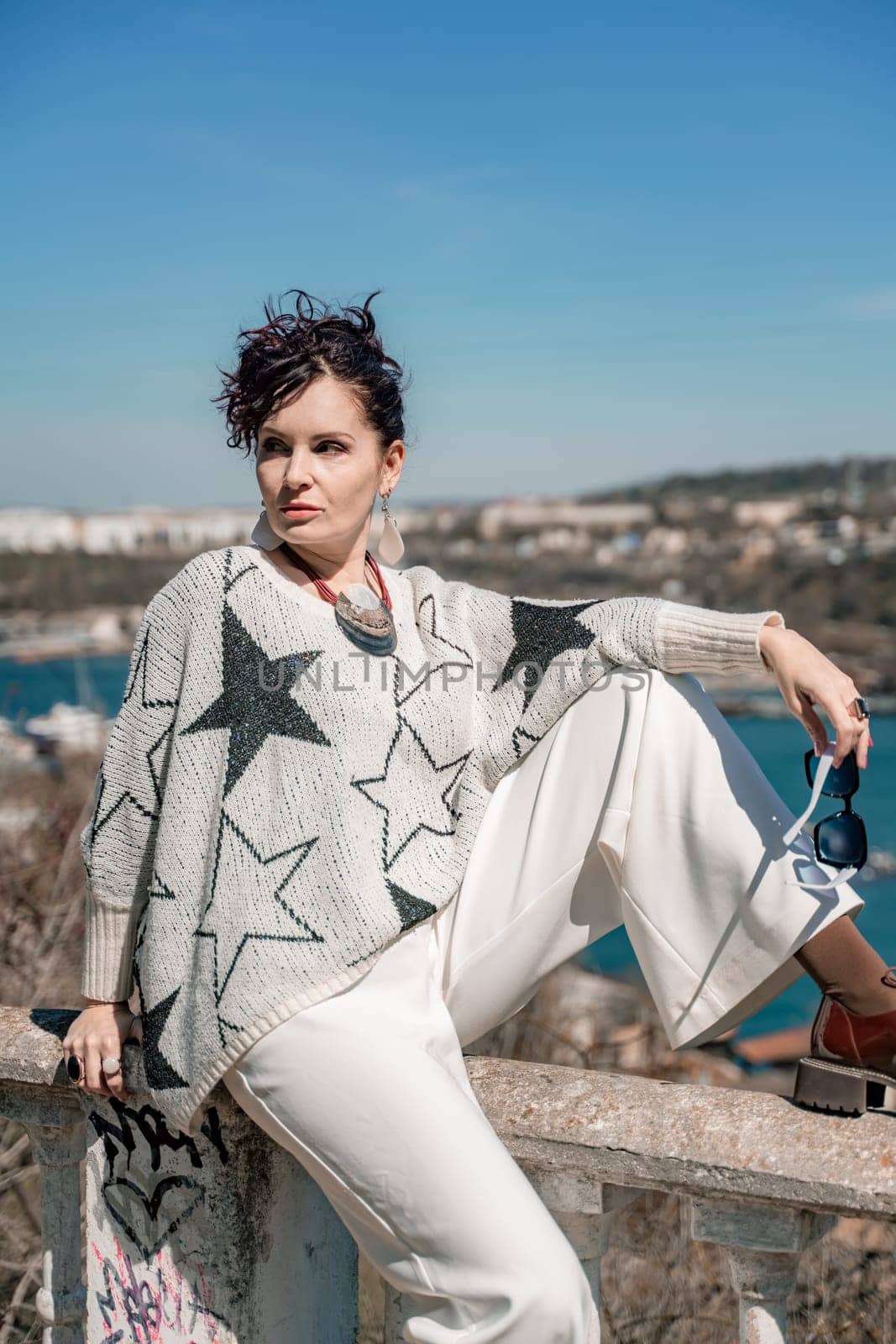 Woman walks around the city, lifestyle. A young beautiful woman in white trousers and a sweater sits on a white fence with balusters and overlooks the sea bay and the city