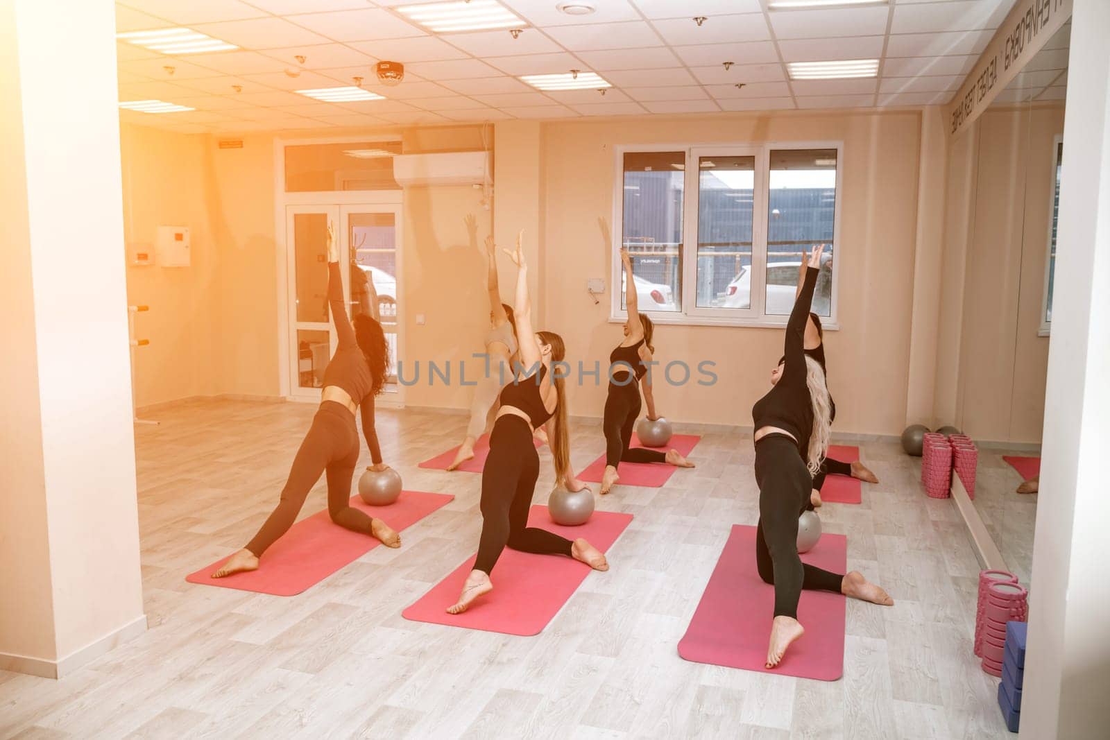 A group of six athletic women doing pilates or yoga on pink mats in front of a window in a beige loft studio interior. Teamwork, good mood and healthy lifestyle concept. by Matiunina