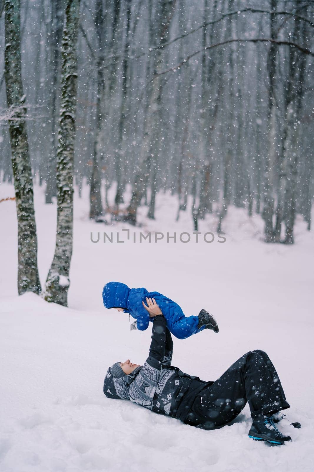 Smiling mother lies in the snow in the forest under a snowfall with her child raised in outstretched arms. High quality photo