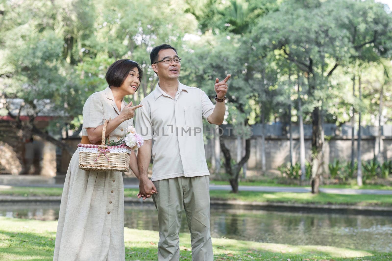 Senior man and wife holding a picnic basket walking among the green trees in the park A retired couple in their 60s walked through the garden together enjoying their vacation. by wichayada