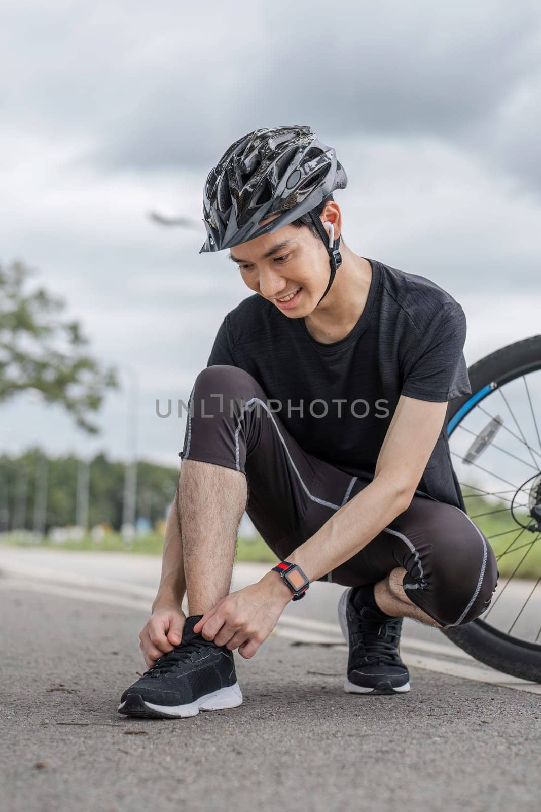 A healthy and strong male cyclist ties his shoe laces before riding a bicycle on a country road. by wichayada