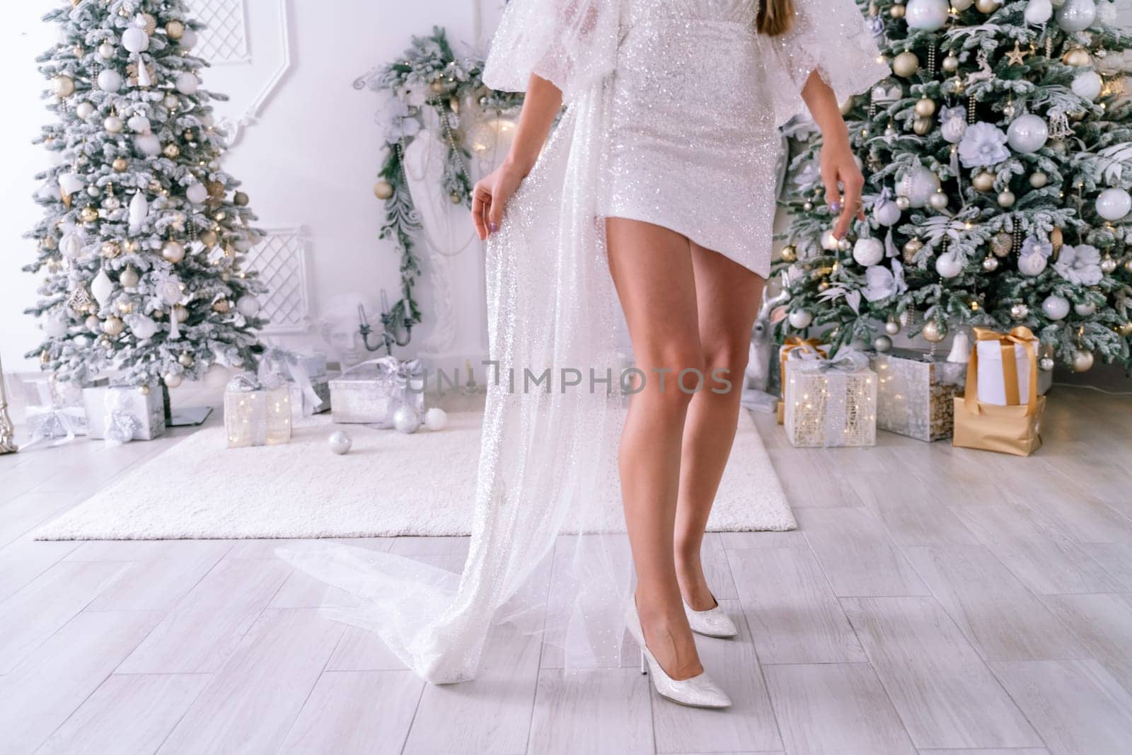 Women's feet christmas tree. Cropped photo of beautiful elegant female legs in white high-heeled shoes and a white short dress, a woman stands against the background of a Christmas tree with gifts.
