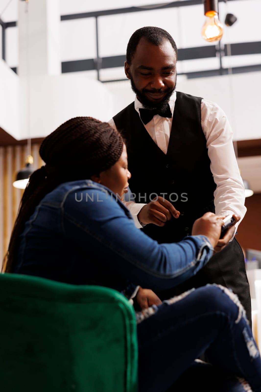 African American woman guest making contactless payment at hotel while resting in lobby. Smiling friendly waiter using NFC enabled card reader to let customer pay for order in restaurant or cafe