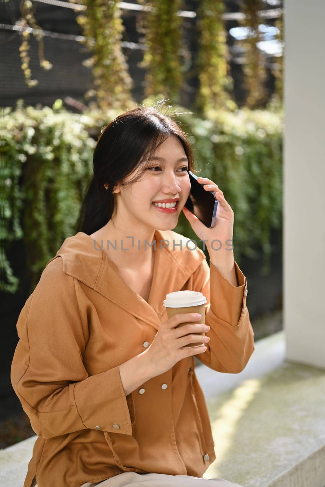 Young businesswoman with take away coffee sitting outside office building talking on mobile phone by prathanchorruangsak