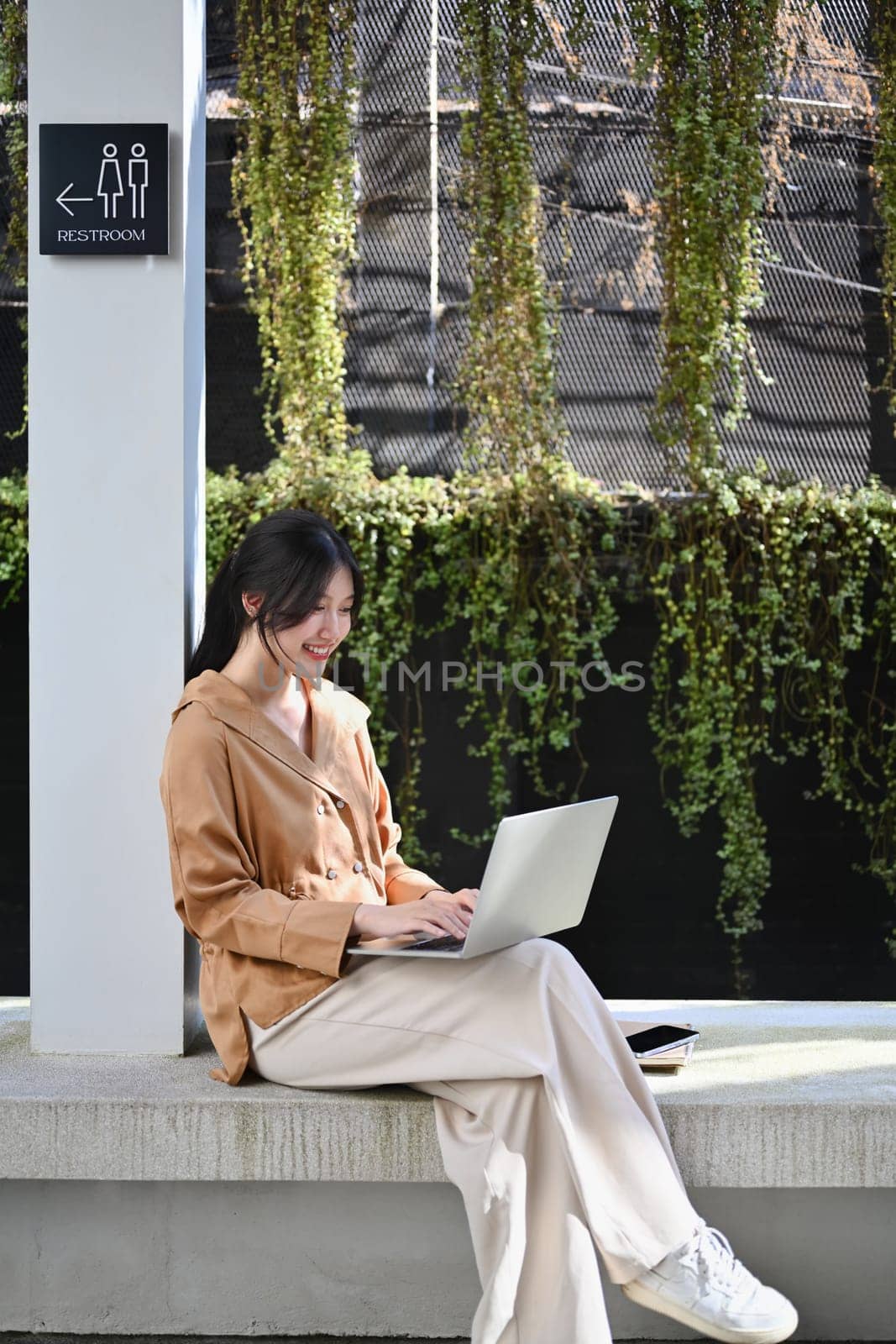 Portrait of happy young businesswoman working with laptop sitting outside her office building by prathanchorruangsak