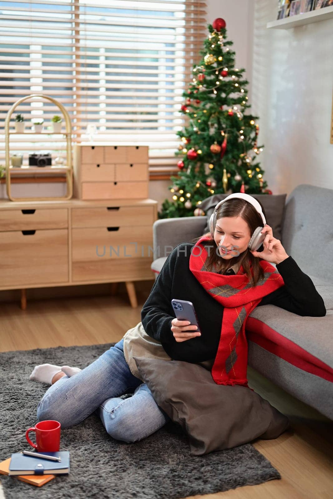 Smiling caucasian woman in headphone using mobile in living room decorated for Christmas.