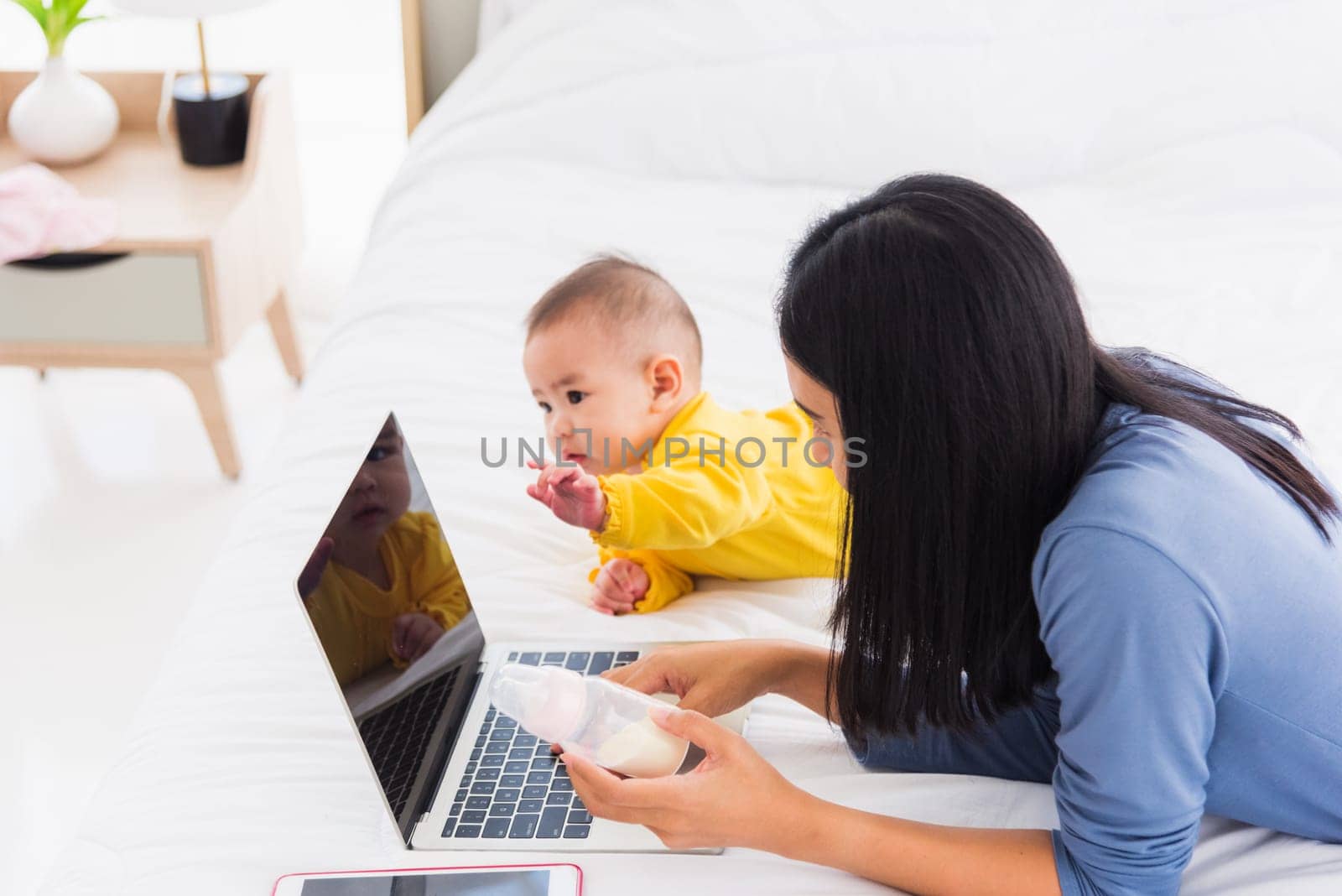 Asian new young beautiful mother finding and reads information on a laptop for milk mixing on a bottle in the bedroom at home