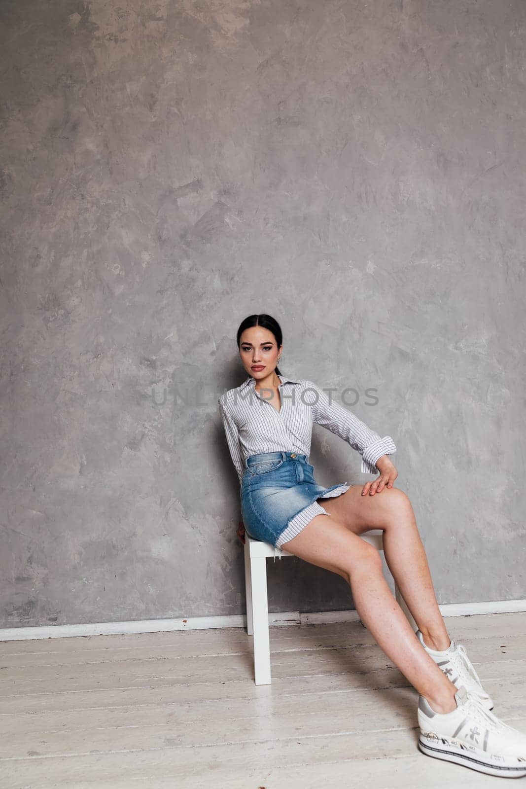 fashionable woman in a denim skirt sits on a chair with a gray background