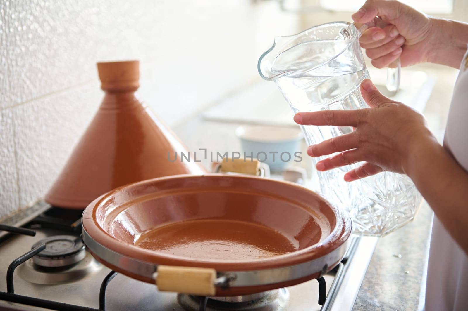 Closeup of an unrecognizable woman, housewife beginning cooking meal in tajine clay pot, pouring some water inside a clay dish standing on a stove. Traditional Moroccan food, culinary and culture