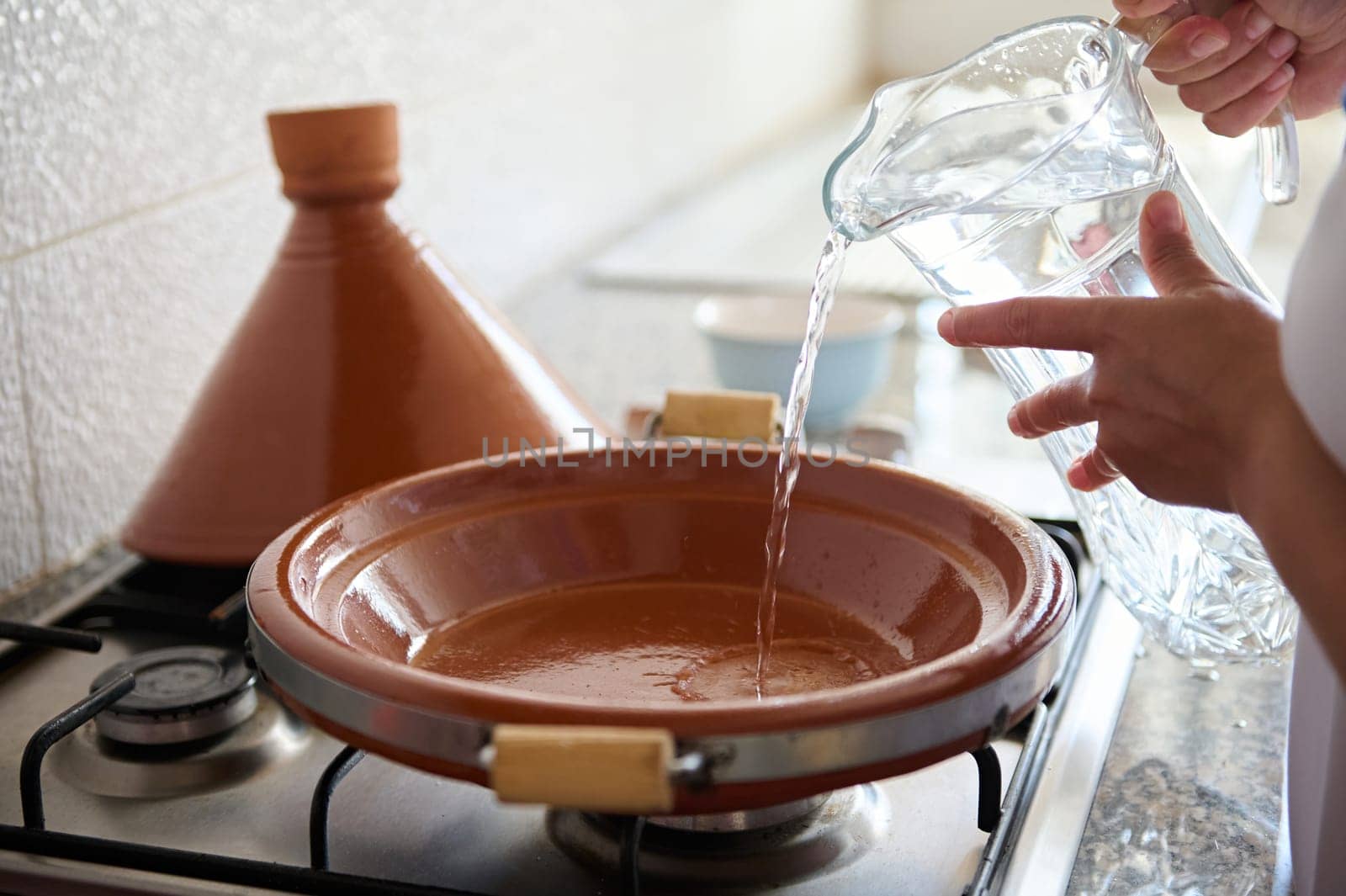 Closeup of an unrecognizable woman housewife pouring some water inside a tagine clay dish, cooking traditional Moroccan meal