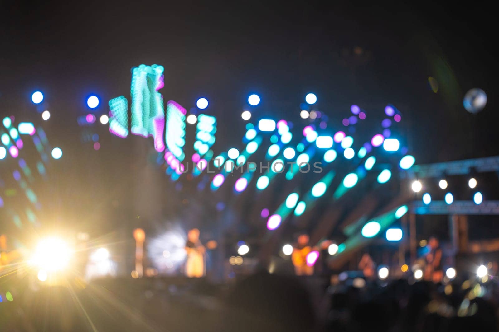 Amid vibrant nightlife a concert festival takes center stage and unrecognizable crowd cheers in front of brilliantly illuminated stage. lens flare captures energy and excitement of music event. by Sorapop