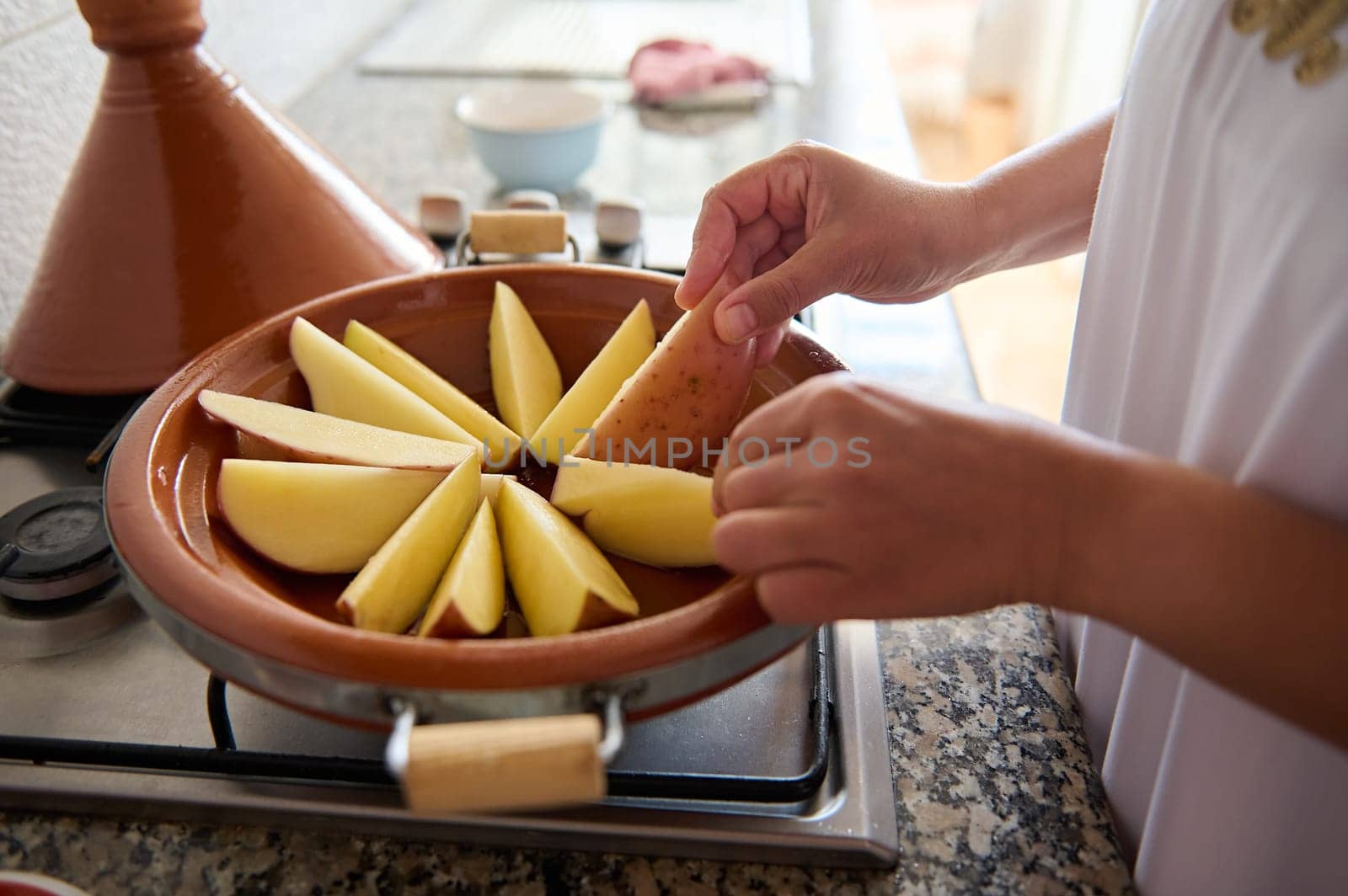 Moroccan tajine clay dish on the stove and Muslim woman housewife stacking slices of organic potato, cooking at home by artgf