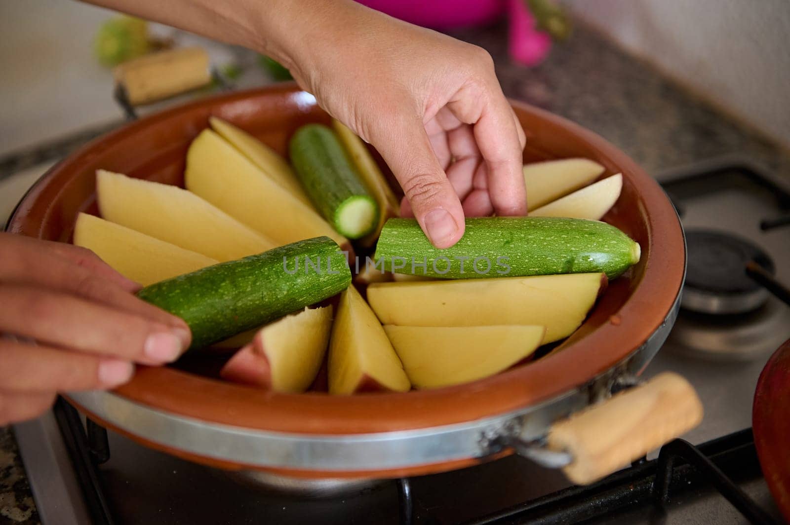 Close-up housewife's hands putting zucchini and potato on the clay dish while cooking Moroccan tagine in the kitchen by artgf