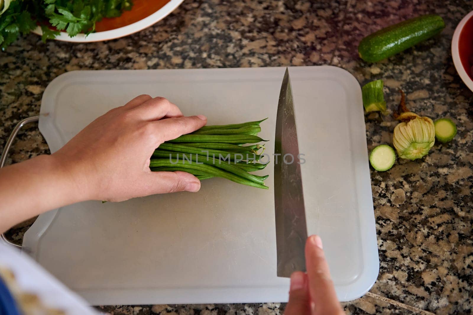 Top view of a housewife, woman chef holding a kitchen knife, chopping fresh green beans on a cutting board, standing by marble kitchen counter in the home kitchen. Raw food. Vegetarianism. Close-up