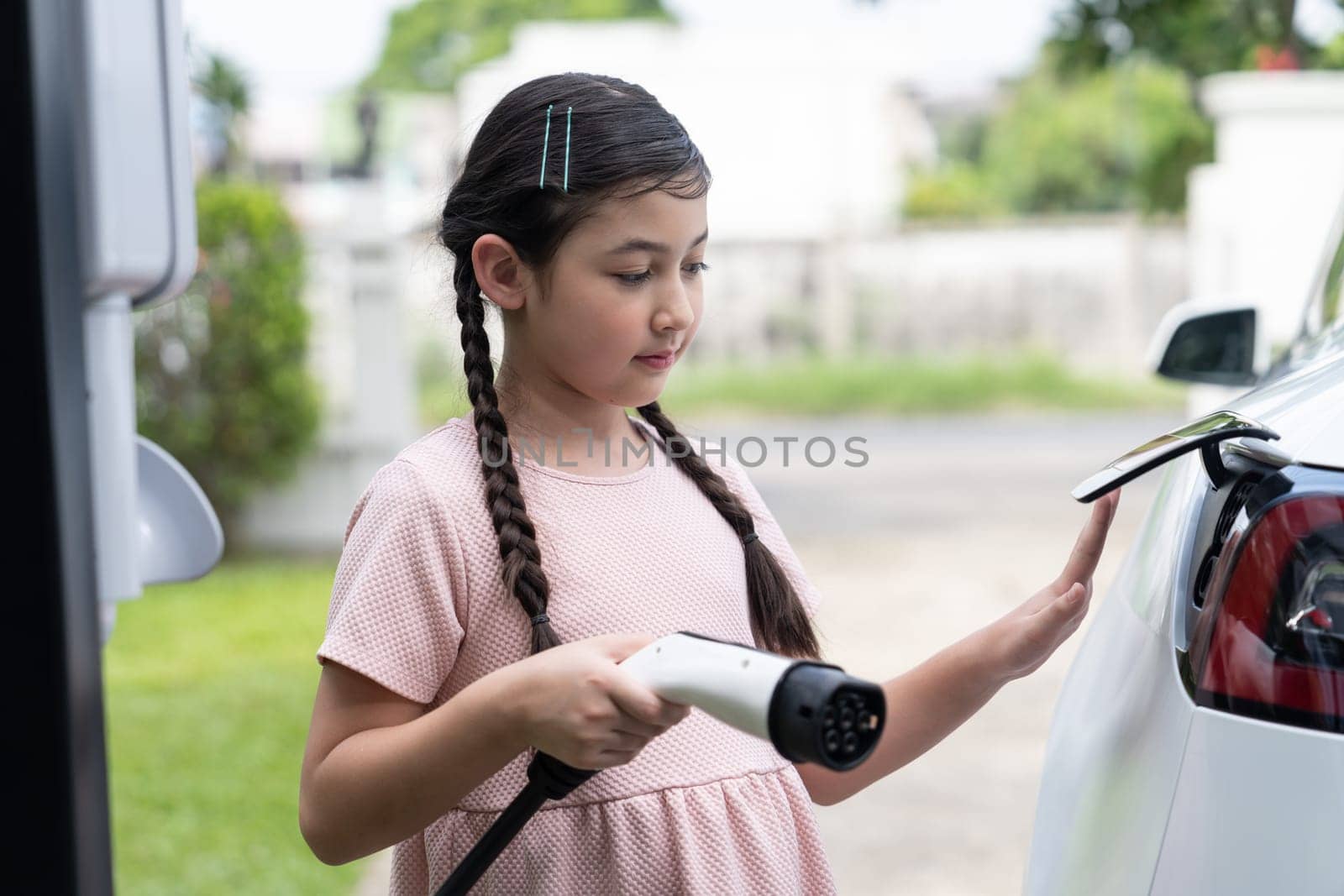 Happy little young girl learn about eco-friendly and energy sustainability as she recharge electric vehicle from home EV charging station. EV car and sustainable future generation concept. Synchronos