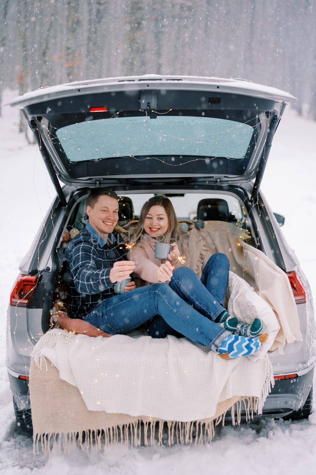 Smiling boyfriend and girlfriend are sitting in the trunk of a car under snowfall with sparklers and mugs of coffee by Nadtochiy