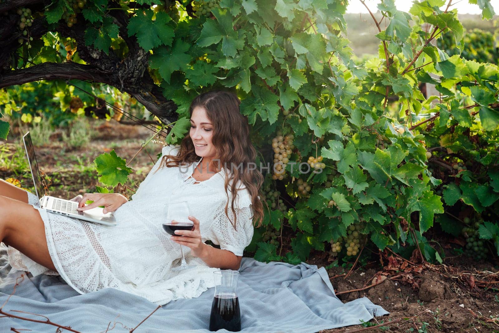 a beautiful woman sits under a grape with a laptop and a glass of red wine