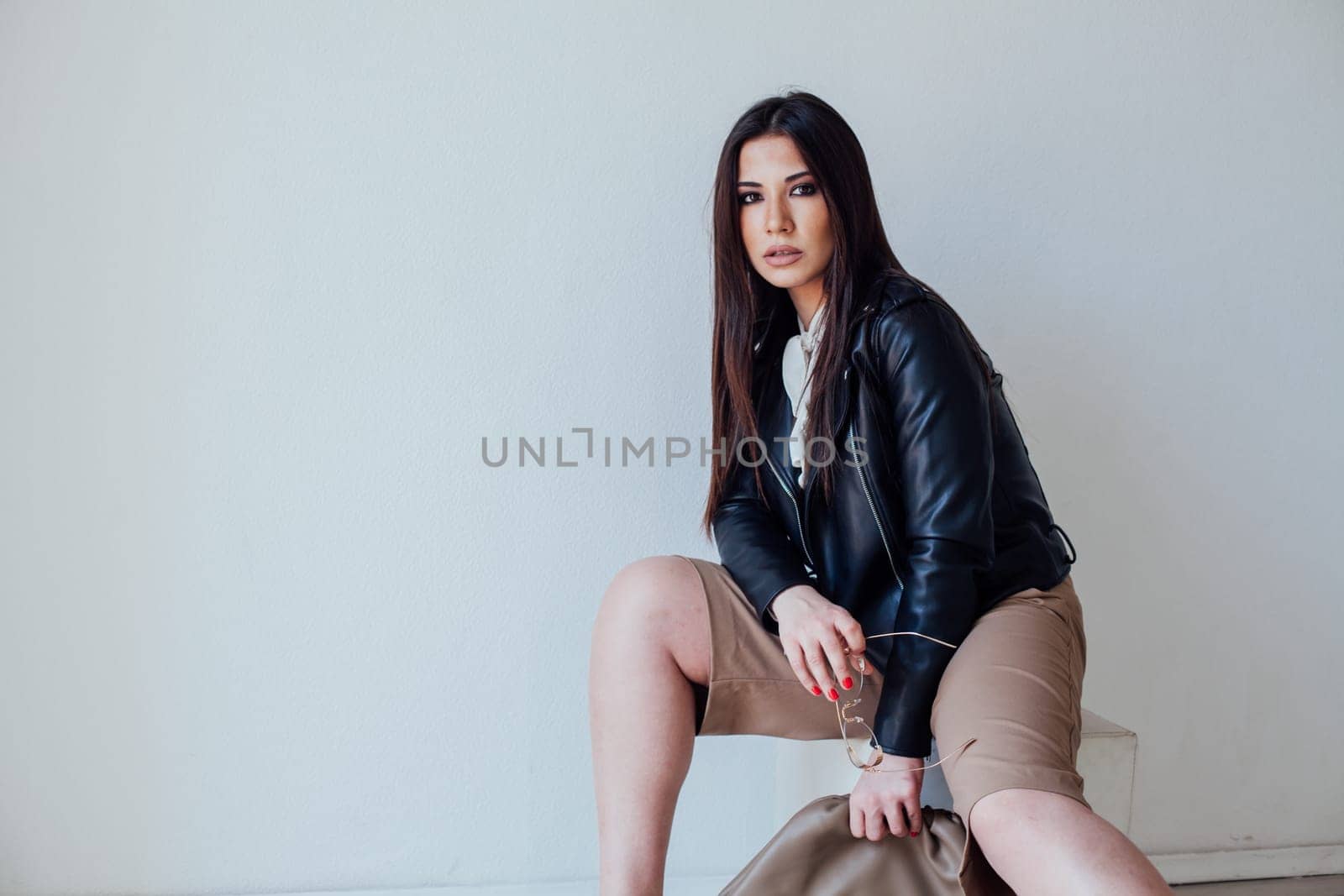 a brunette woman in a leather jacket sits on a low chair by Simakov
