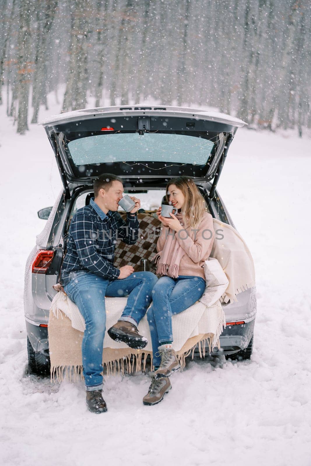 Couple drinking coffee from cups while sitting in the trunk of a car in a snowy forest by Nadtochiy