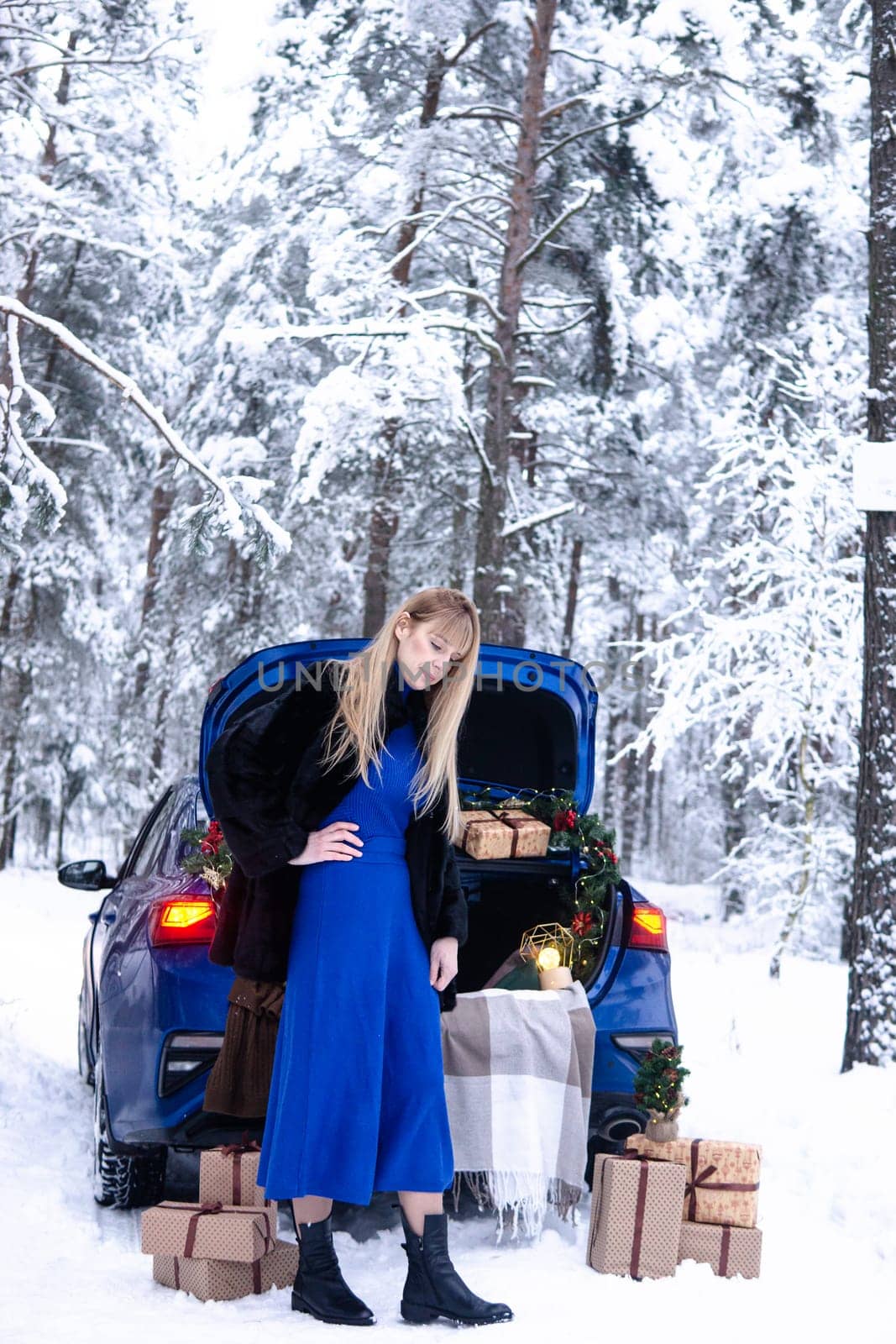 Woman in winter snowy forest in blue dress next to blue car decorated with Christmas decor. Christmas and winter holidays concept