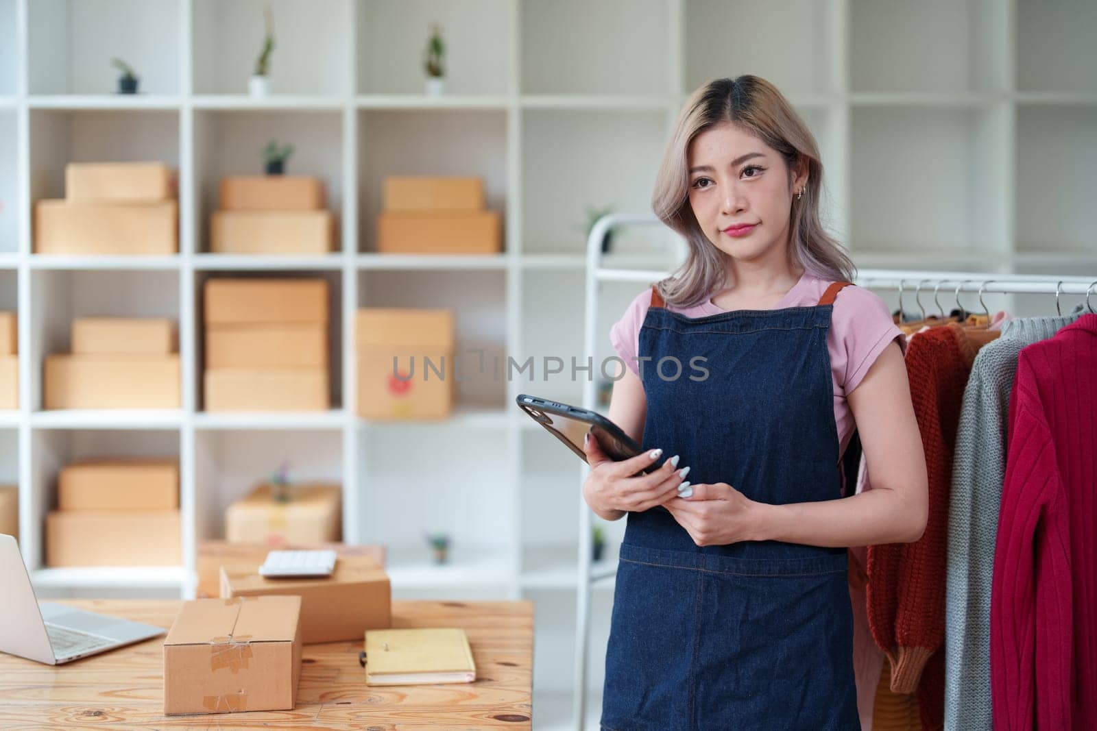 Portrait Of Asian Female Owner Of Fashion Store Checking Stock In Clothing Store With using notebook successful happy smile at small business, sme or ecommerce concepts by Manastrong