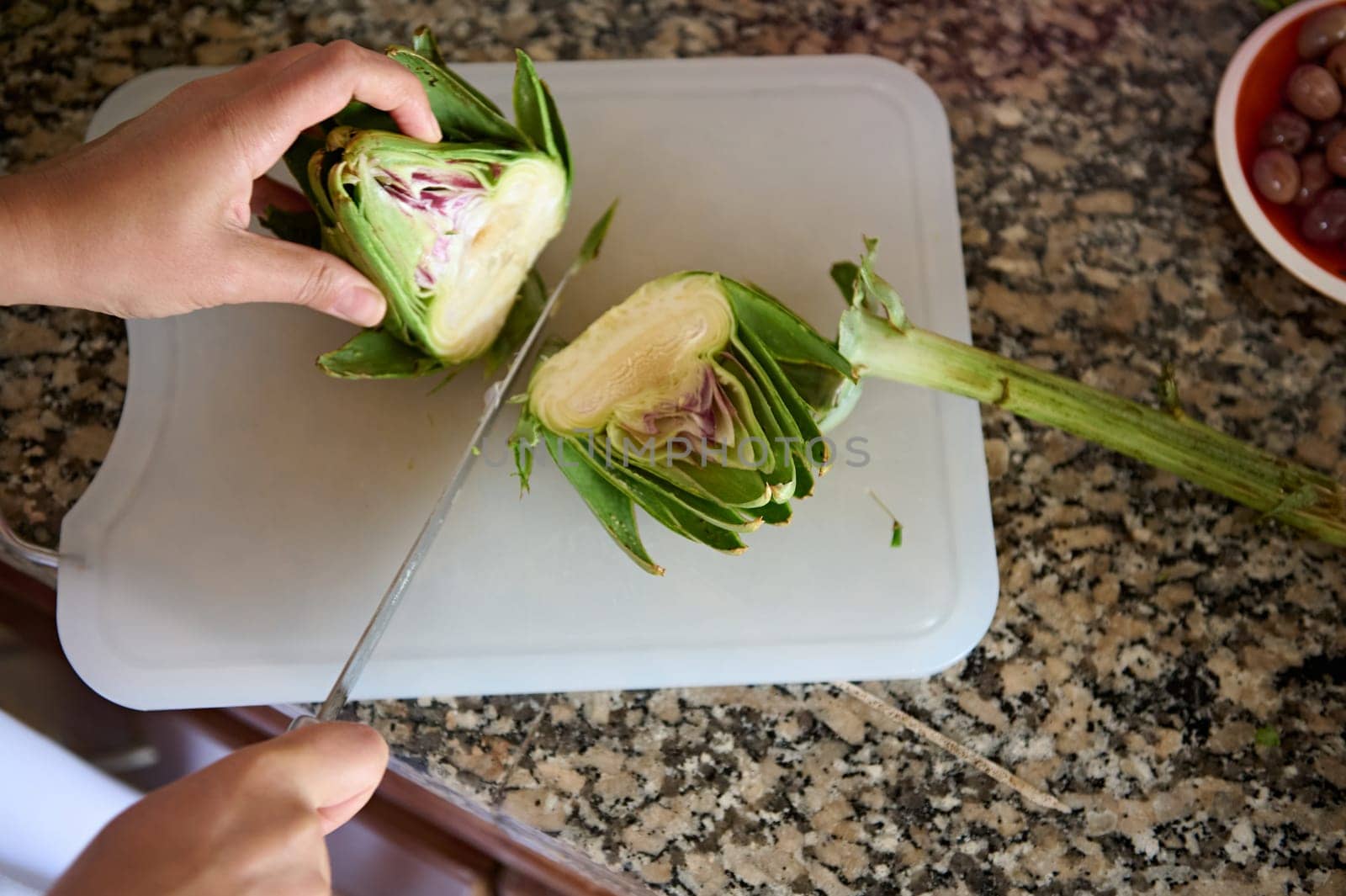 Top view chef hands cutting artichoke flower on a board. Two halves of fresh ripe organic artichoke on cutting board by artgf