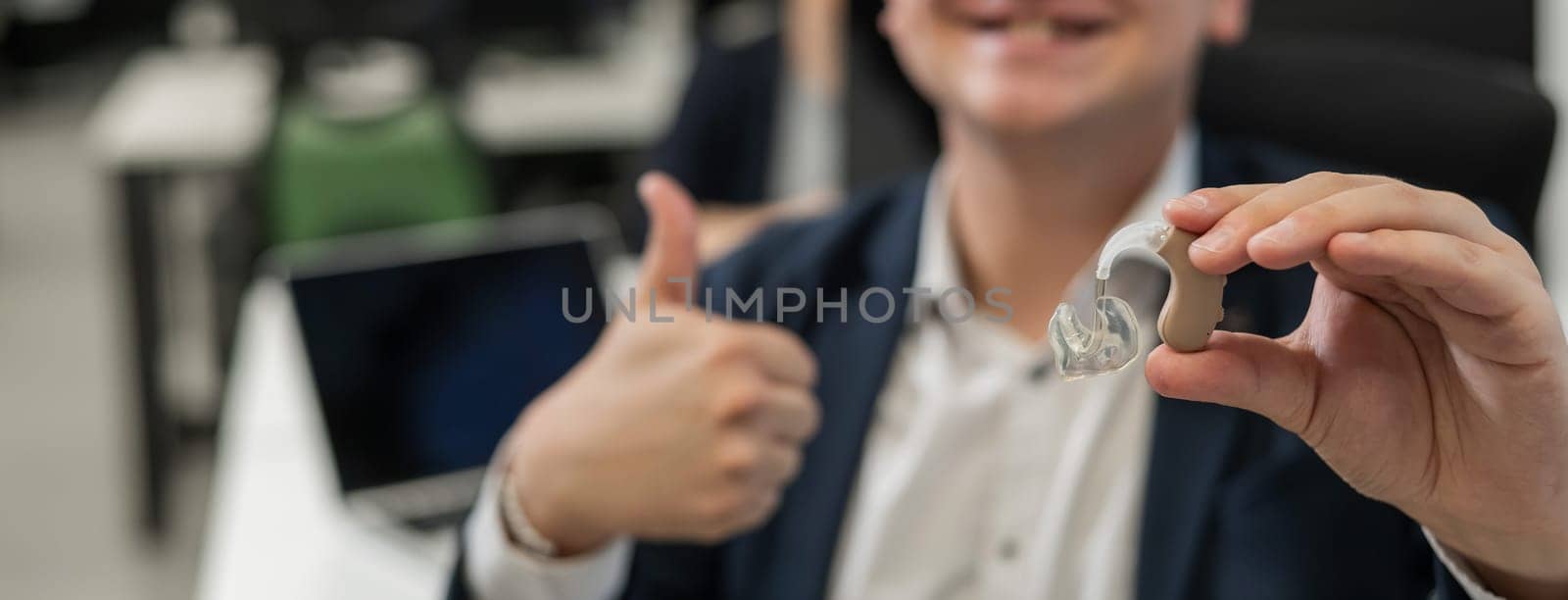 Portrait of a man holding a hearing aid and showing thumbs up in the office. Widescreen