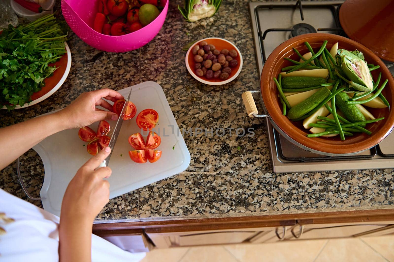 Top view of a housewife, woman chef holding a kitchen knife, chopping fresh juicy tomatoes on a cutting board, standing by marble kitchen counter in the home kitchen. Raw food. Vegetarianism. Close-up