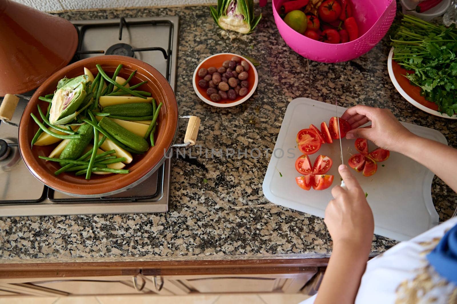 Close-up view of a housewife, woman chef cook holding a kitchen knife, slicing fresh red tomatoes on a cutting board, standing by marble kitchen counter in the home kitchen. Raw food. Vegetarianism.