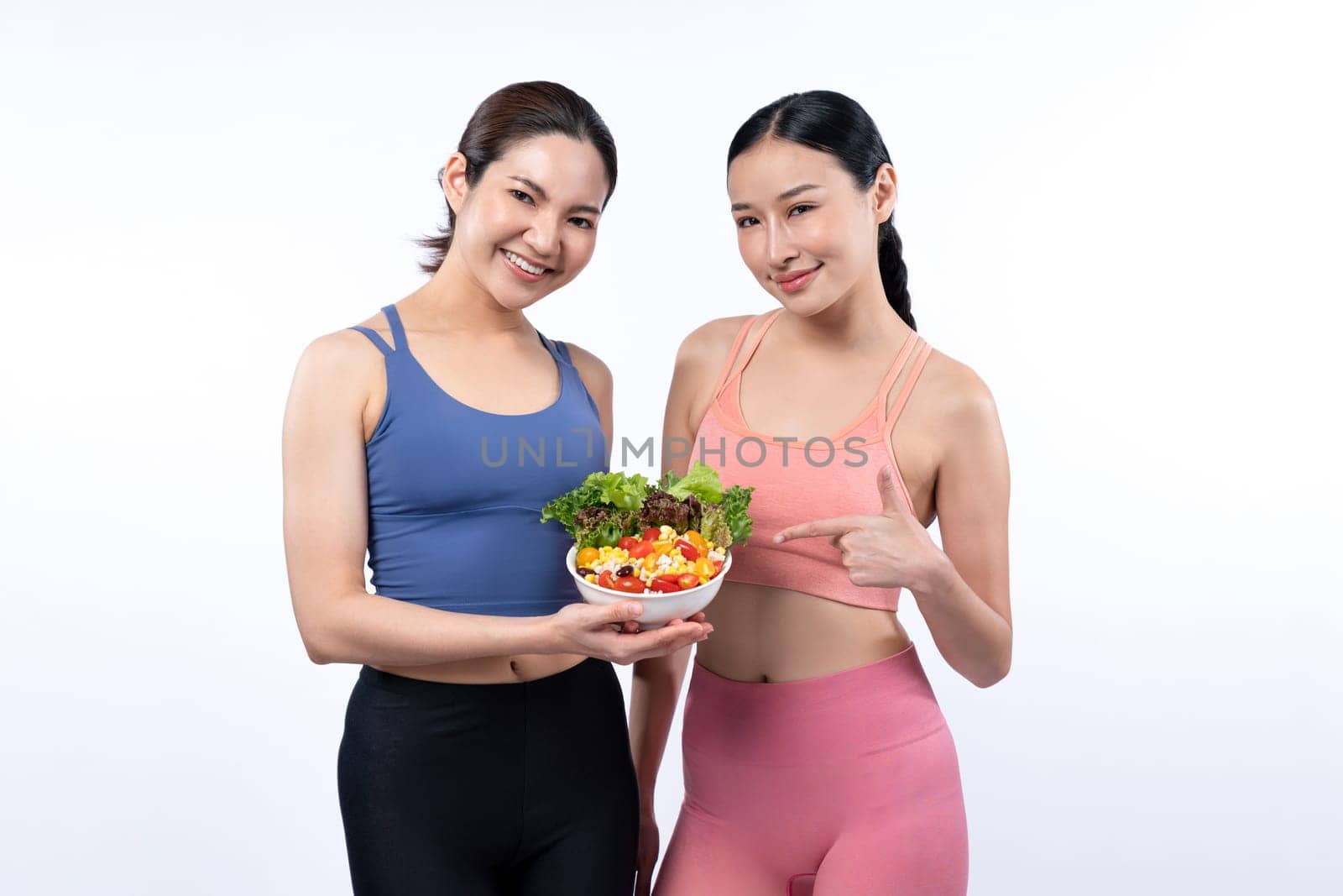 Two young sporty Asian women in sportswear holding salad bowl fill with fruit and vegetable. Natural youthful and fit body lifestyle people with balance nutrition on isolated background. Vigorous