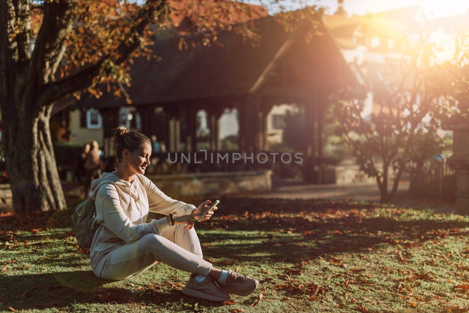 Young Fashionable Teenage Girl With Smartphone In Europian Park In Autumn Sitting At Smiling. Trendy Young Woman In Fall In Park Texting. Retouched, Vibrant Colors. Beautiful Blonde Teenage Girl Wearing Casual Modern Autumn Outfit Sitting In Park In Autumn. Retouched, Vibrant Colors, Brownish Tones. by Andrii_Ko