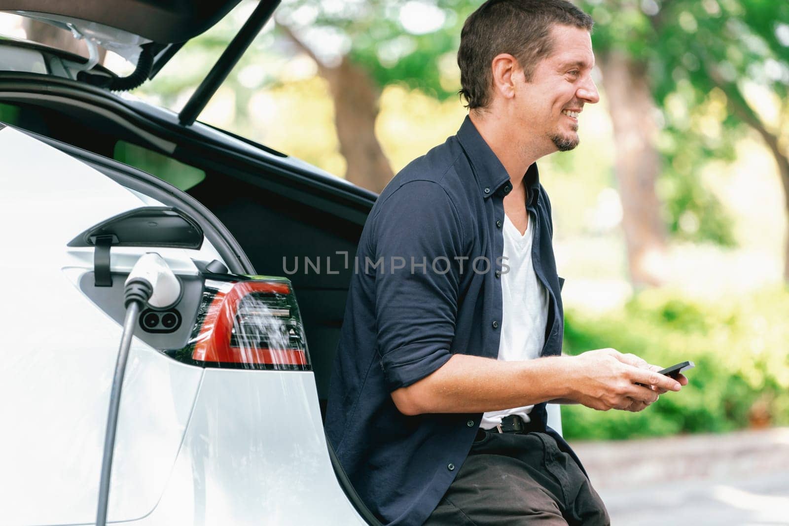 Man using smartphone online banking application to pay for electric car battery charging from EV charging station during vacation holiday road trip at national park or summer forest. Exalt