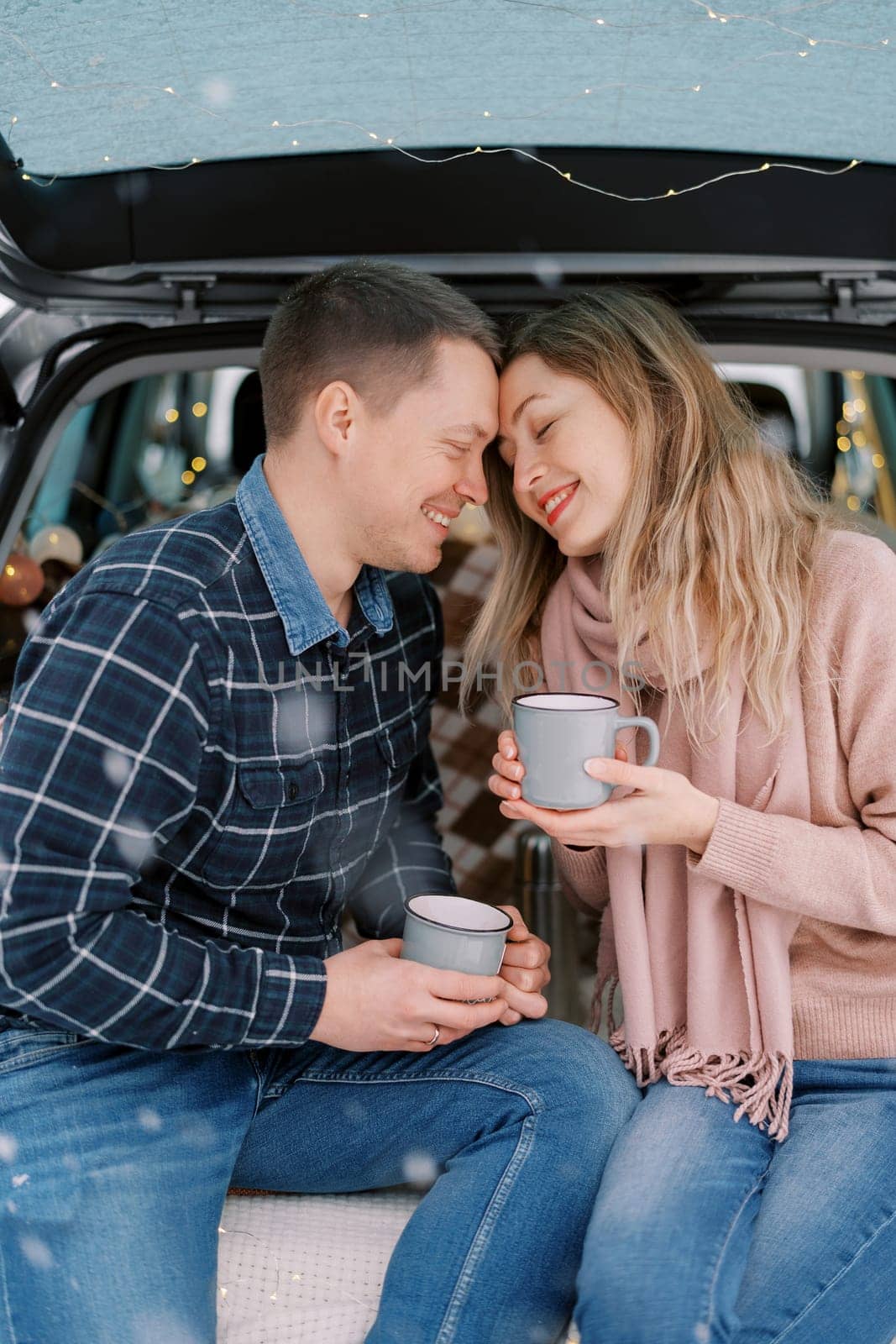 Smiling man and woman touching foreheads while sitting with cups in car trunk. High quality photo