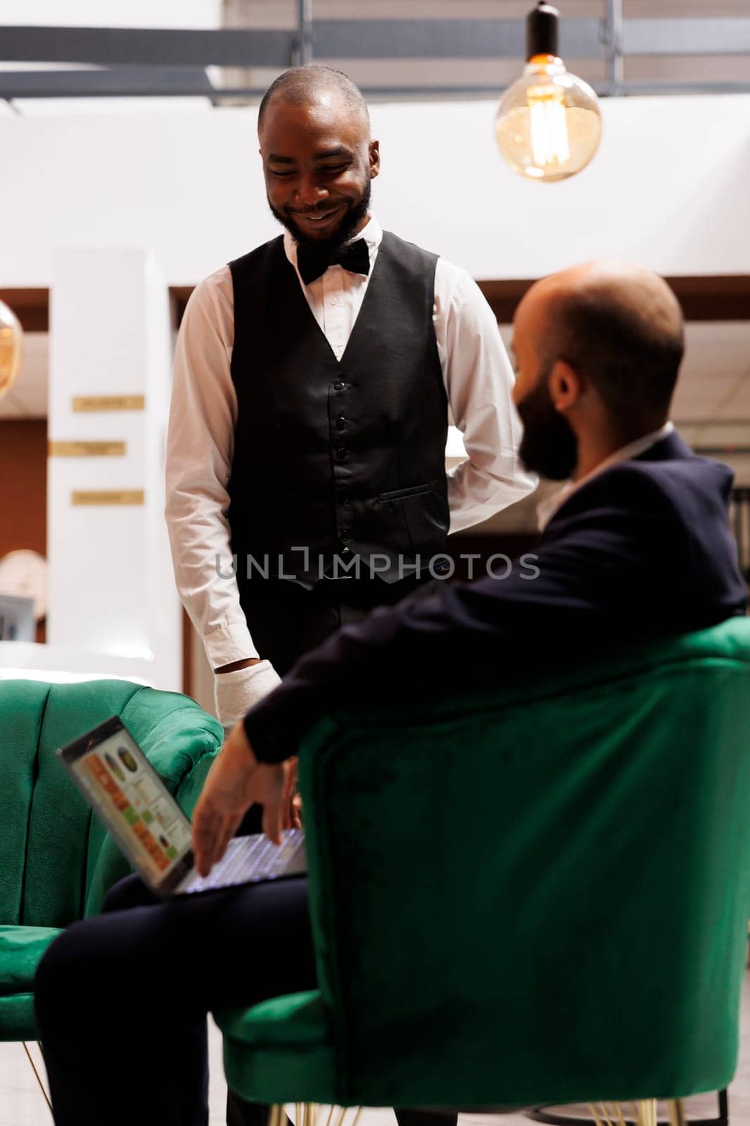 Bellboy talking to client in lounge area, assisting with luxury services while he waits to do check in procedure. Businessman using laptop for official project partnership, business trip.