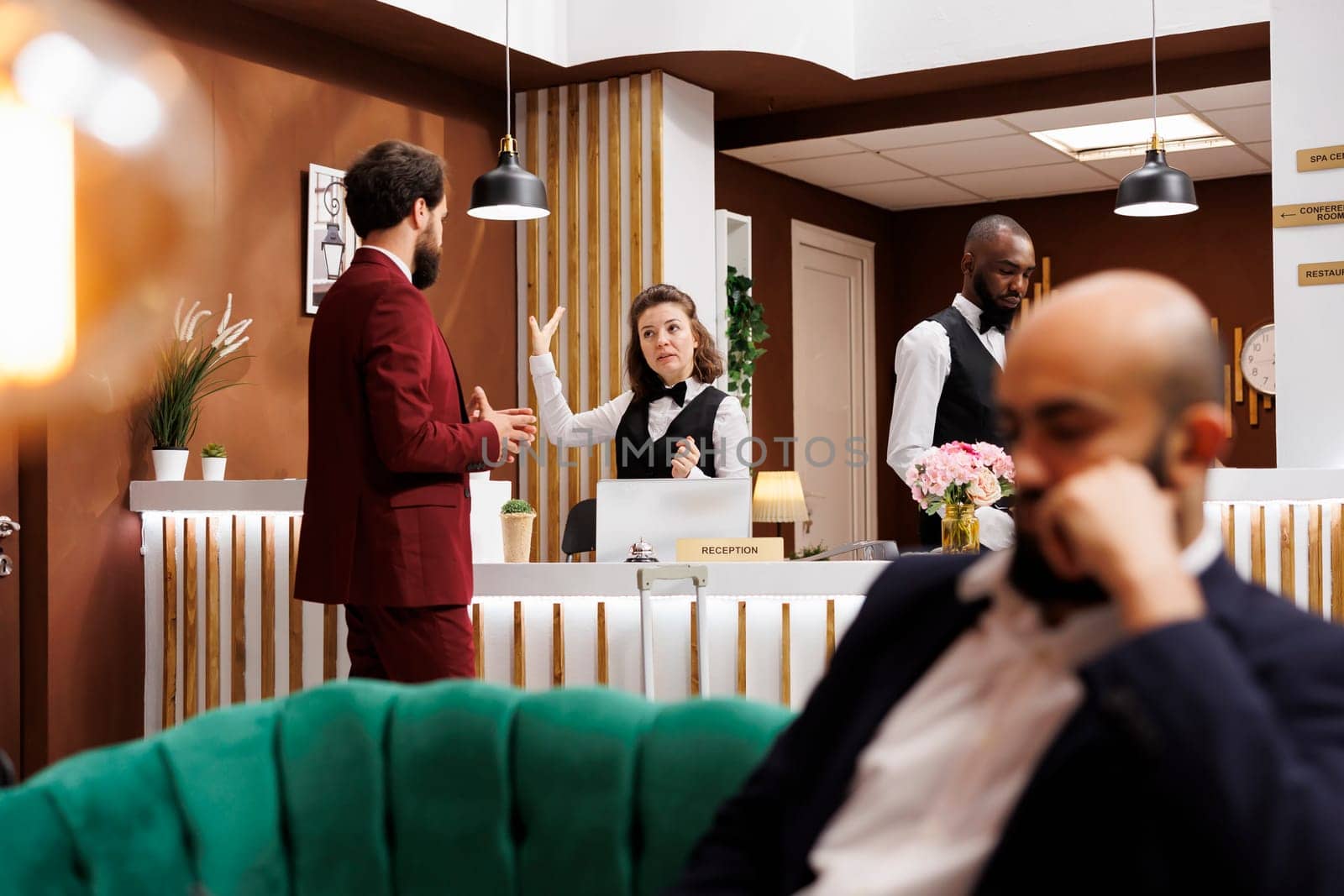 Businessman chats with front desk staff in reception lobby, asking about hotel room services after doing check in. Formal guest arriving at resort, travelling on business trip.