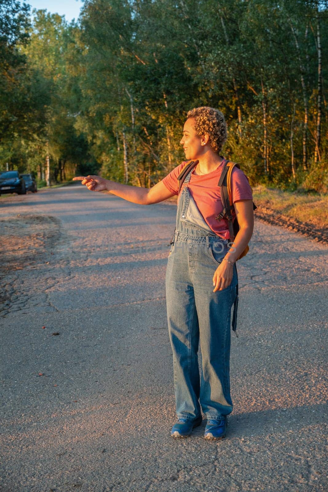 Brazilian millennial girl in denim overalls stands on a forest road and points with her hand at sunset, golden hour by KaterinaDalemans