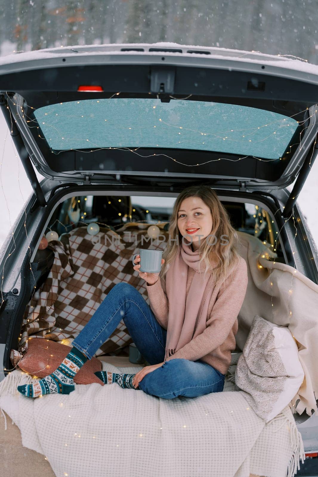 Smiling girl with a mug of tea sits on a blanket in the trunk of a car under snowfall in the forest by Nadtochiy