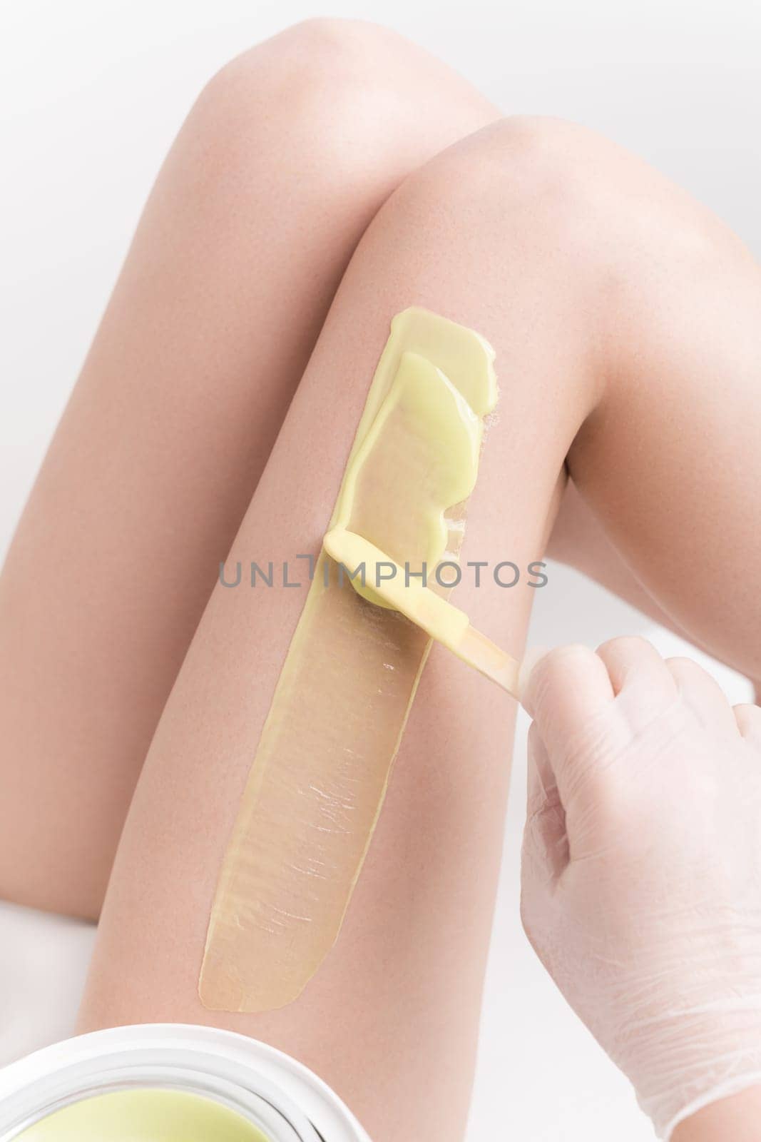 Beautician in gloves applying green hot wax on slim woman leg using spatula. Depilation with hot wax in beauty salon. Part of photo series.