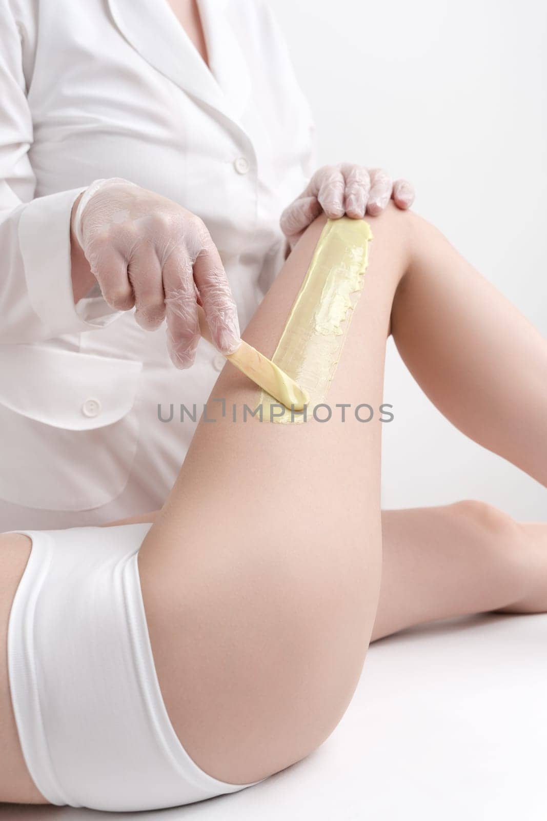 Unrecognizable beautician in white coat applying green hot wax on woman leg using spatula by Alexander-Piragis