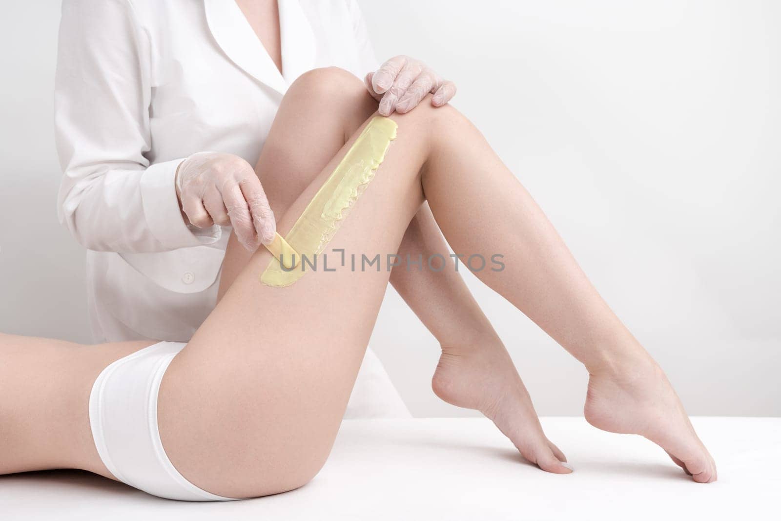Cosmetologist applying green hot wax on slim woman leg using spatula while woman lying down on couch by Alexander-Piragis