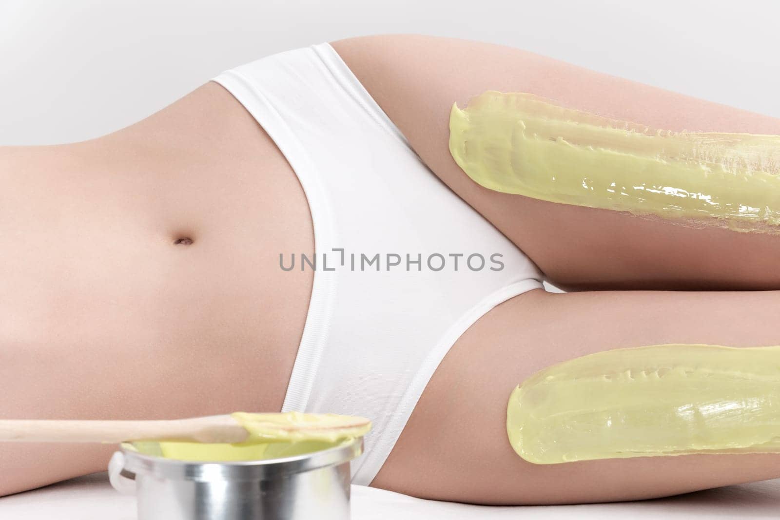 Legs of slim woman while she lying down on couch during depilation process with green hot wax in professional beauty salon. Healthy slim female body in white panties. Part of photo series.