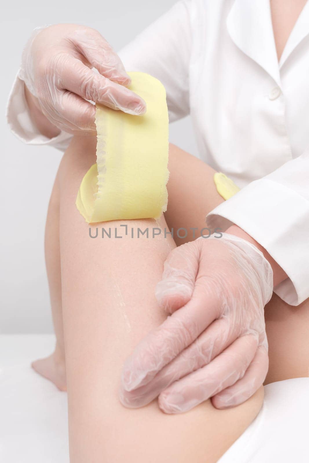 Depilation procedure - cosmetologist hands in gloves removing hair on women leg. Waxing process with green hot wax in professional beauty salon. Part of photo series.