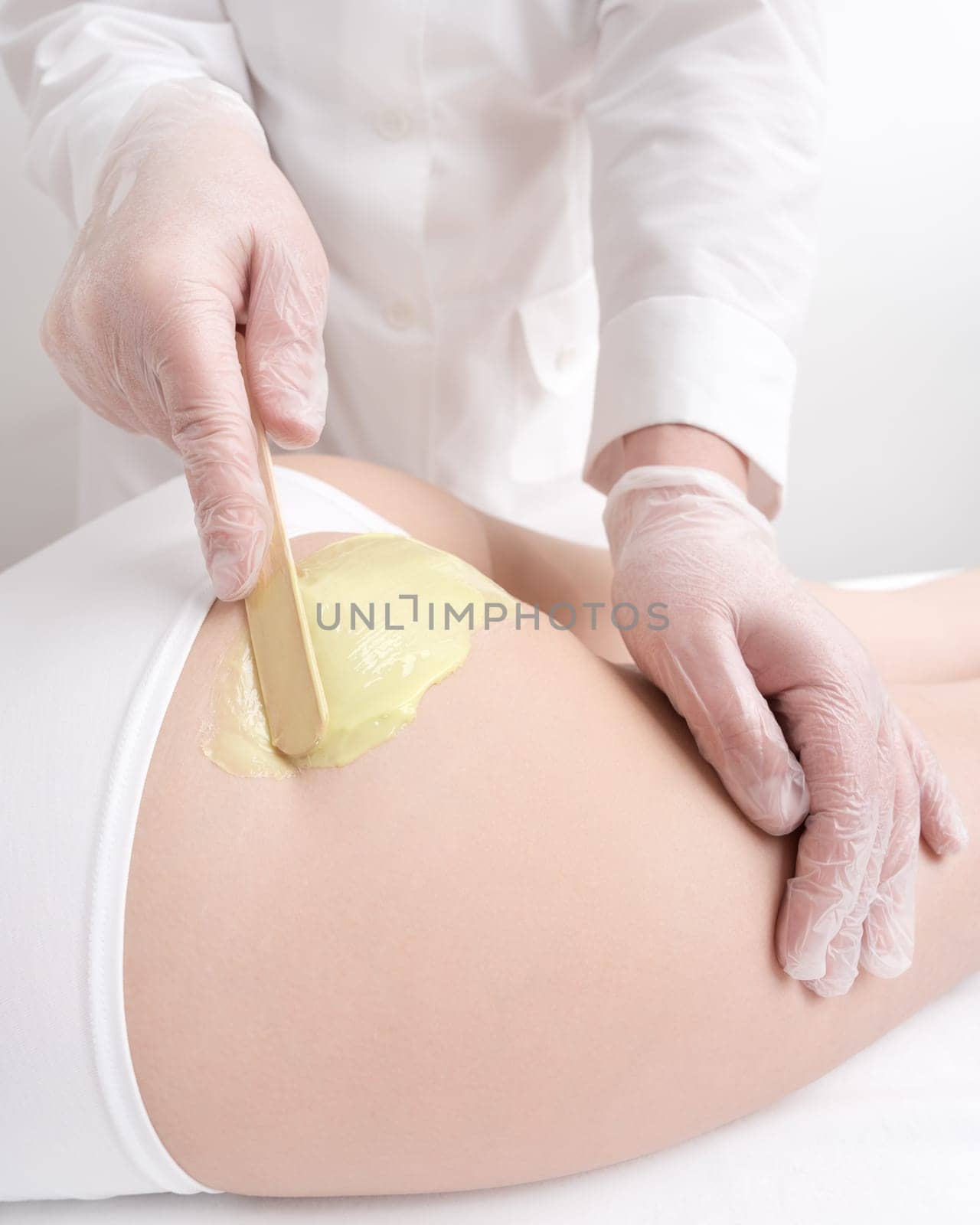 Closeup hands in gloves applies green hot wax on buttocks of woman using spatula. Waxing procedure, depilation with hot wax in professional beauty salon. Part of photo series.