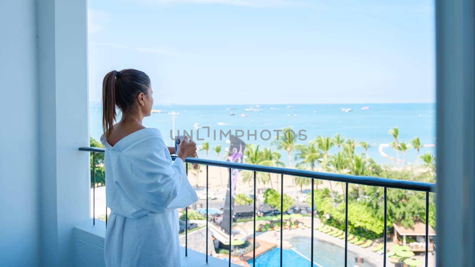 a Asian woman on vacation drinking coffee in the morning looking out the window over the beach and ocean during a luxury holiday