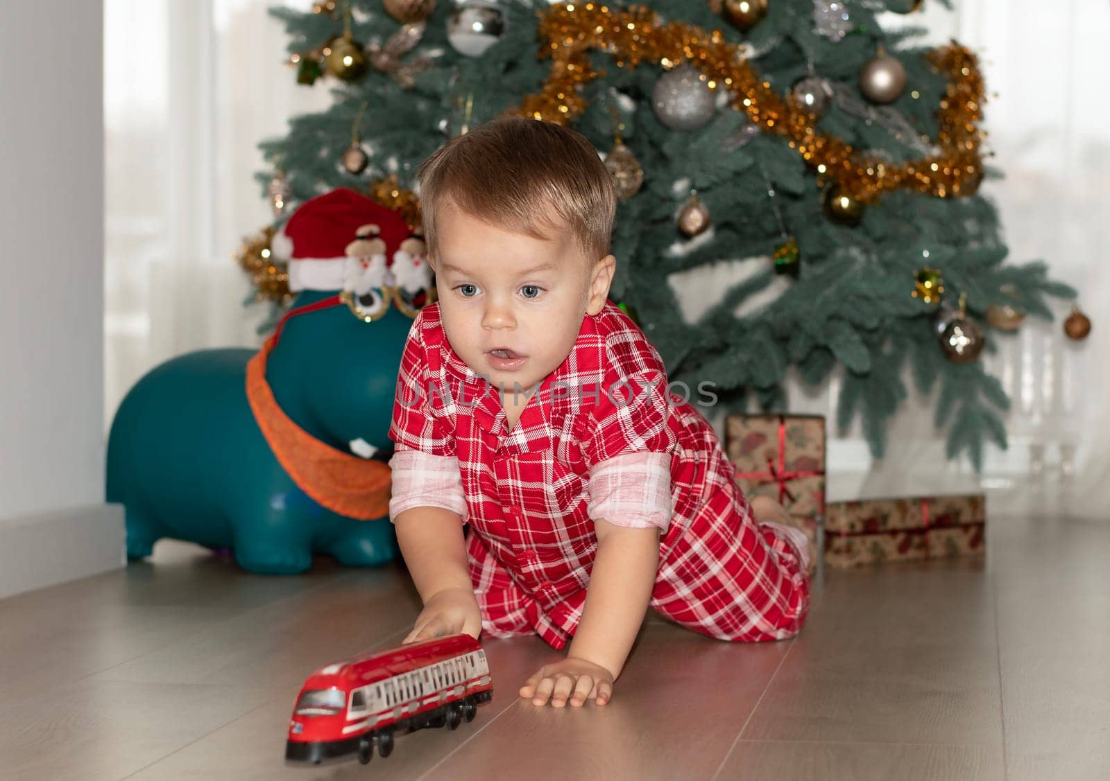 Christmas concept. New Year 2024. A small handsome boy in red checkered pajamas sits on the floor and plays with a red steam locomotive against the background of a decorated Christmas tree with gifts. Close-up.