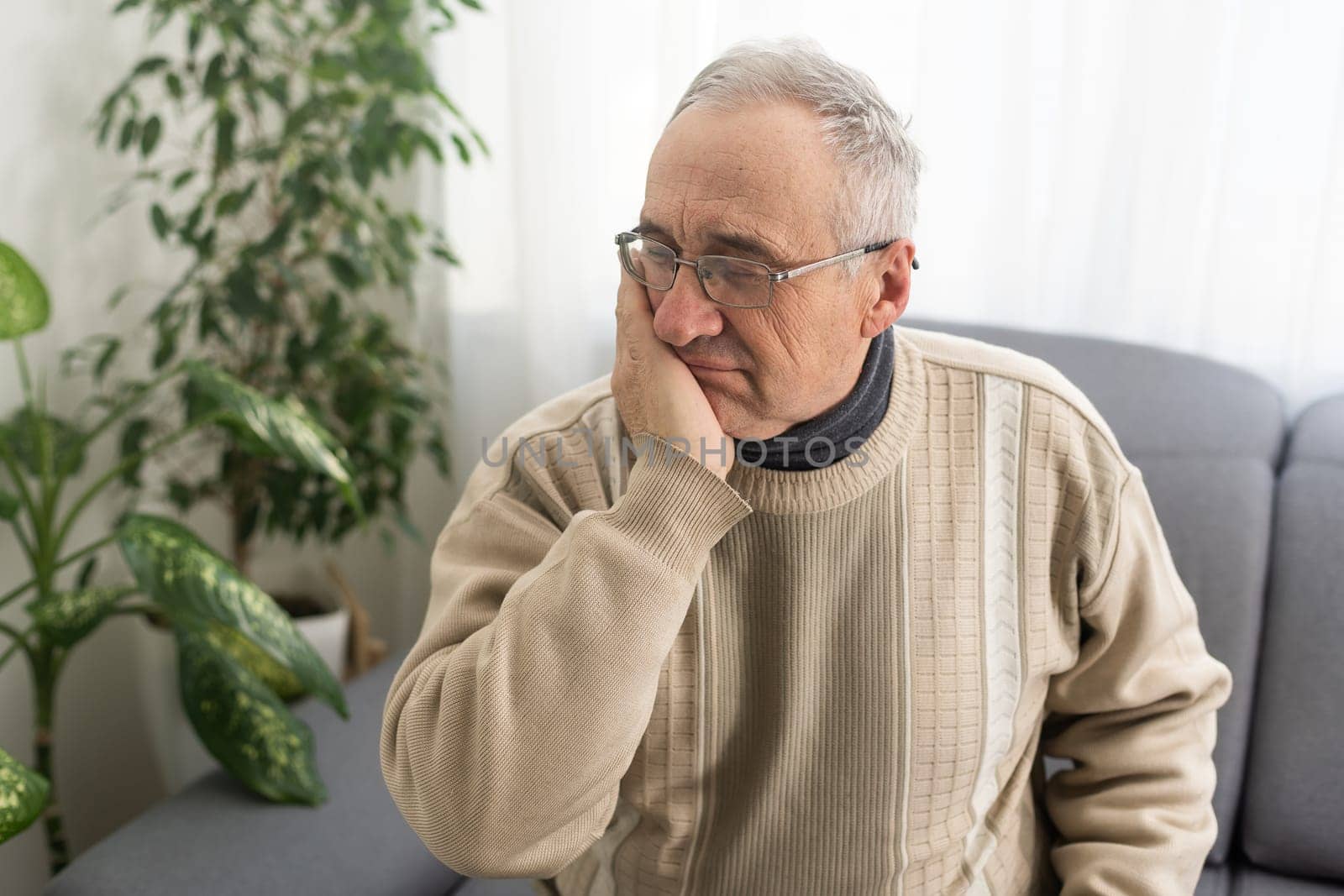 Old man with toothache. Elderly senior man has toothache. Unhappy man face in tooth pain sitting on sofa at home, feel sick unwell. Sad aged man hand holding his chin. Adult suffering toothache.