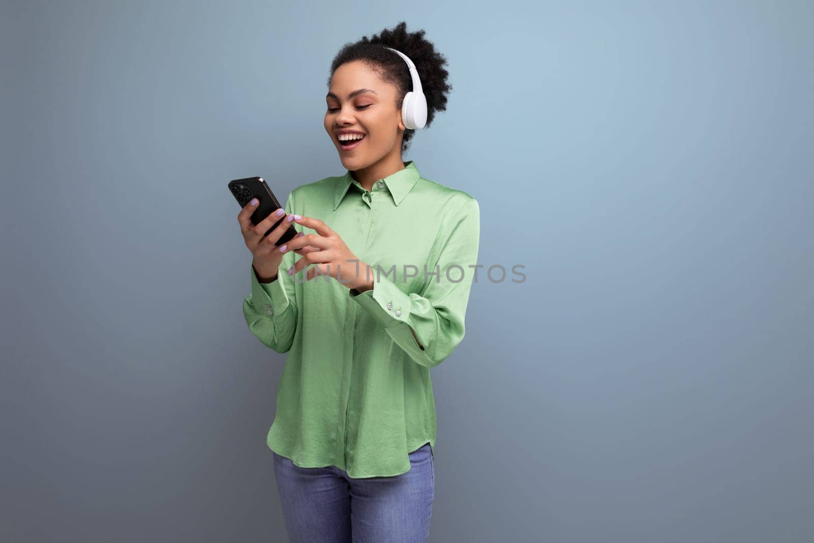 young cheerful positive hispanic brunette lady dressed in a green stylish blouse is watching a video on a smartphone using headphones for sound on the background with copy space.